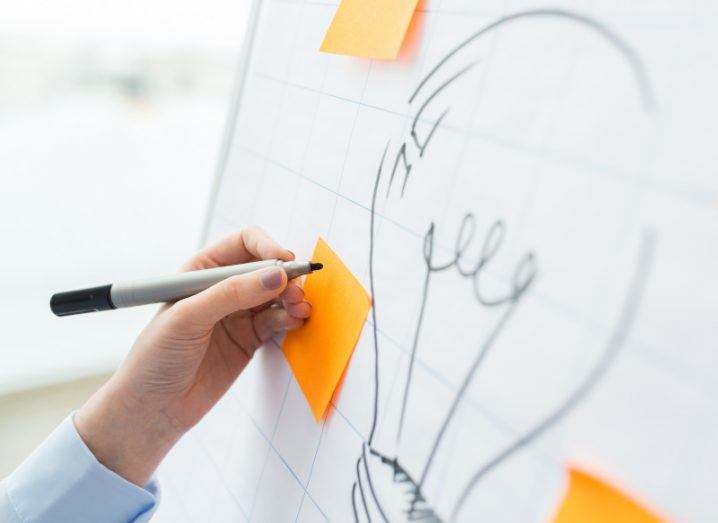 Close-up image of a hand drawing on an orange post-it note, which is stuck onto a white board with a drawing of a lightbulb.