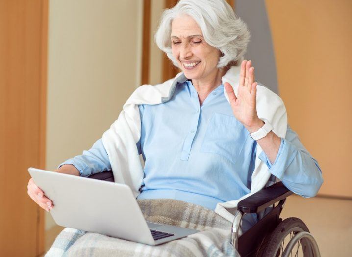 Woman in a wheelchair wearing a blue shirt and white cardigan, waving at a laptop screen.