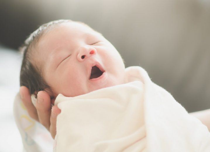 Newborn baby yawning while being held by an adult, against a white and grey background.