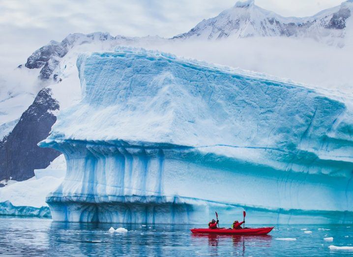 Two people in a red kayak paddling past a large glacier at Antarctica.