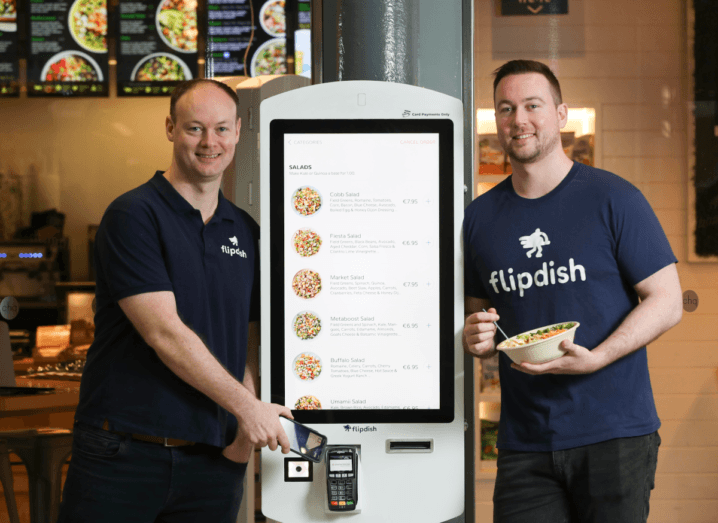 Two men standing on either side of a white kiosk with a touch screen for ordering food. They are both wearing navy T-shirts that say 'Flipdish'.