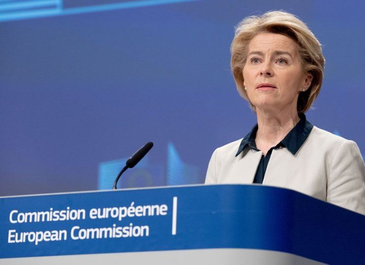Ursula von der Leyen in a white suit jacket speaking at a podium on behalf of the European Commission.