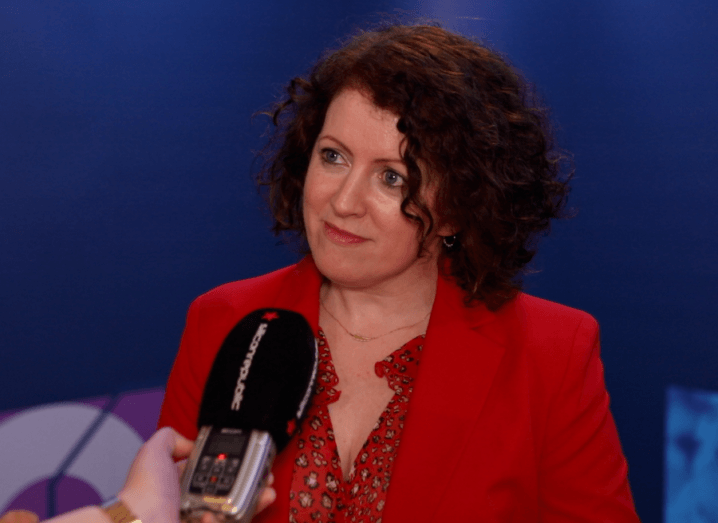 A woman in a red blazer and a red blouse with short, curly brown hair stands in front of a blue background.