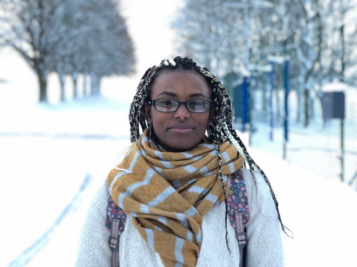 A woman wearing a scarf and a carrying a backpack stands in a corridor of trees in a snowy park.