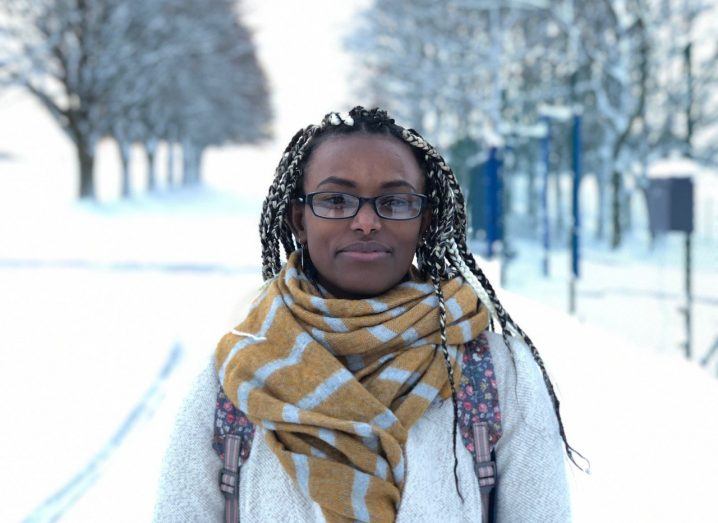 A woman wearing a scarf and carrying a backpack stands in a corridor of trees in a snowy park. She is Abeba Birhane.