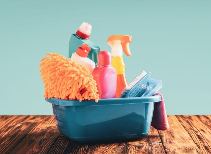 A blue basin filled with an assortment of household cleaning products on a wooden surface against a blue background.