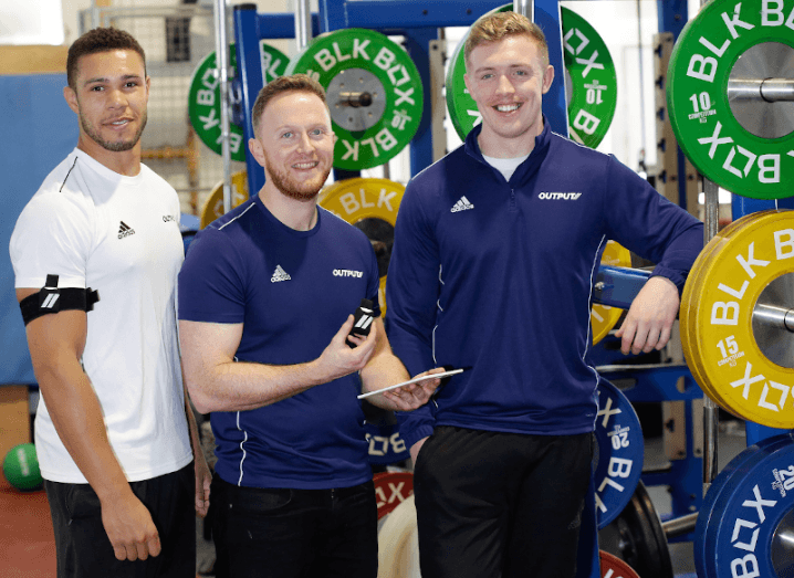Three men in sports shirts that read 'Output' stand in a gym, while one is holding a small tech device.