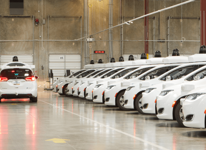 A row of white Waymo cars in a warehouse, with one car driving down the middle.