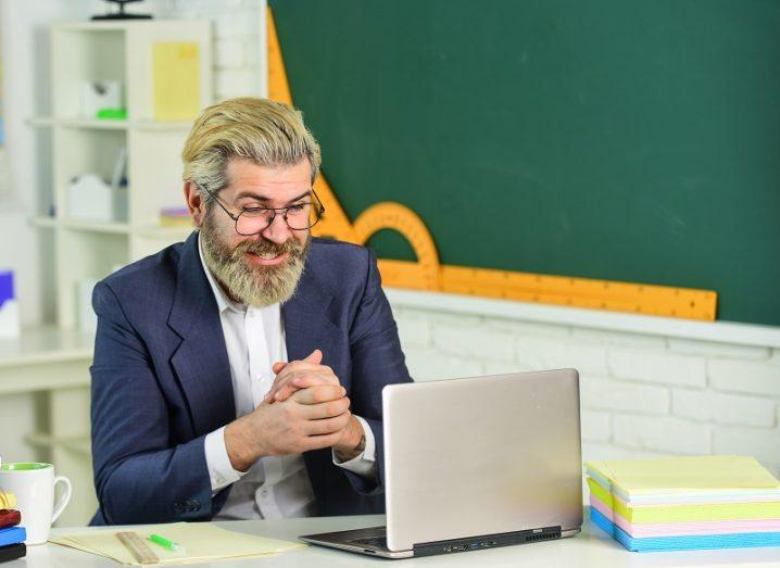 Bearded man smiling in a school classroom talking to someone on a laptop screen.