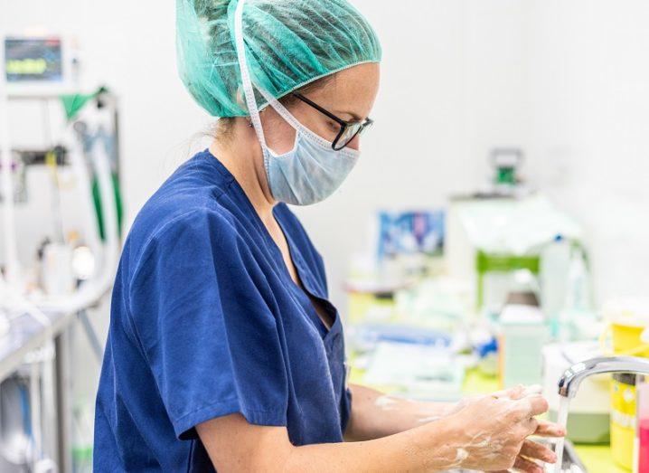 A healthcare worker wearing scrubs and a face mask washes their hands at a sink in a bright white room.