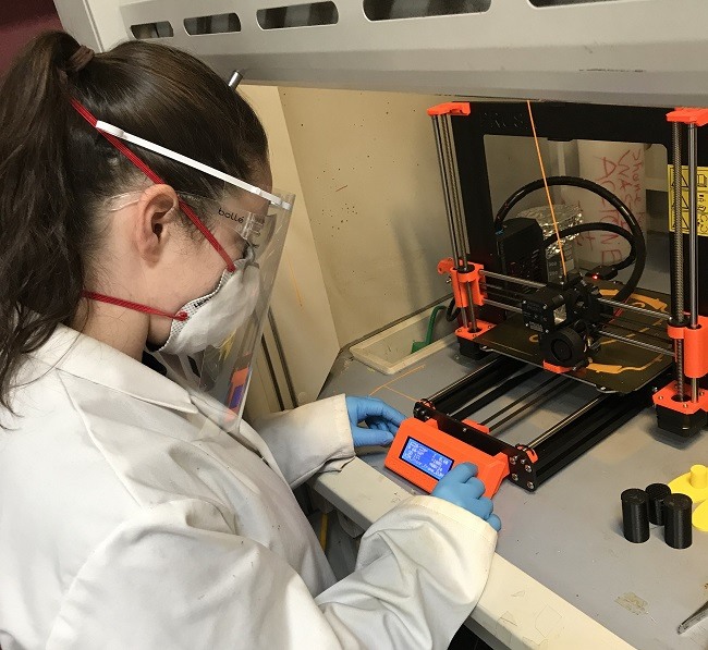 Heather O'Conner wearing a face shield and white lab coat while operating a 3D printer.