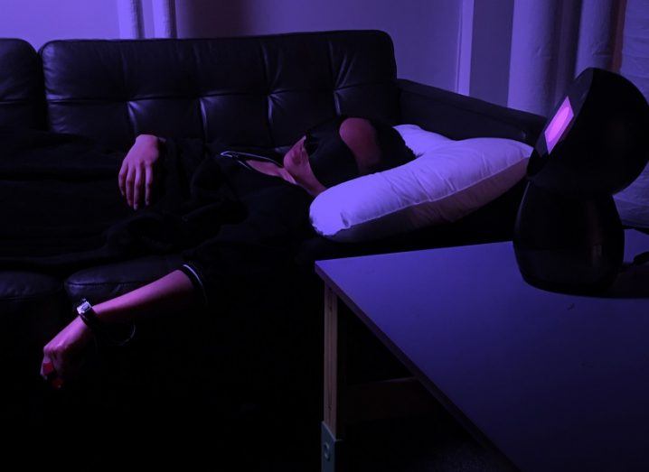 Woman using the Dormio device while asleep on the couch in a dark room lit by a fluorescent purple light.