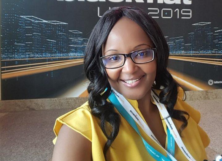 A head and shoulders photo of Noureen Njoroge, a smiling woman with long black hair, wearing glasses and a yellow blouse.