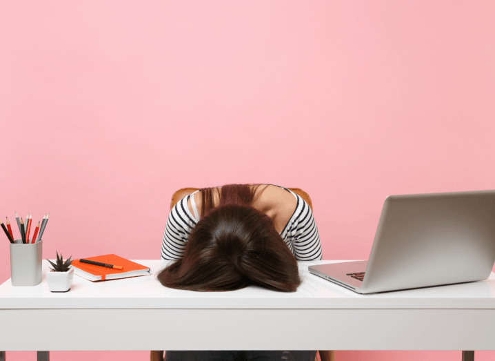 A frustrated woman with her head on a desk in front of a pink background.