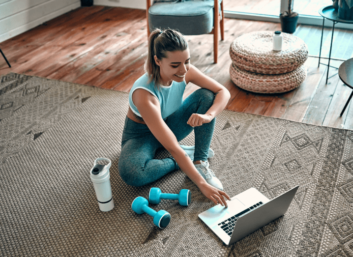 A woman wearing gym gear sitting on the floor with a bottle of water and dumbells in front of a laptop.