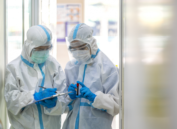 Two medical staff wearing personal protective equipment in a hospital setting. One is holding a vial of blood and the other is writing on a chart.