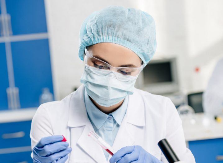 A close-up of a female scientist in a lab coat wearing a facemask, hair net and gloves, holding a test tube.