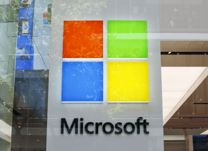 The Microsoft logo behind a pane of glass, with reflections of leaves on the outside.