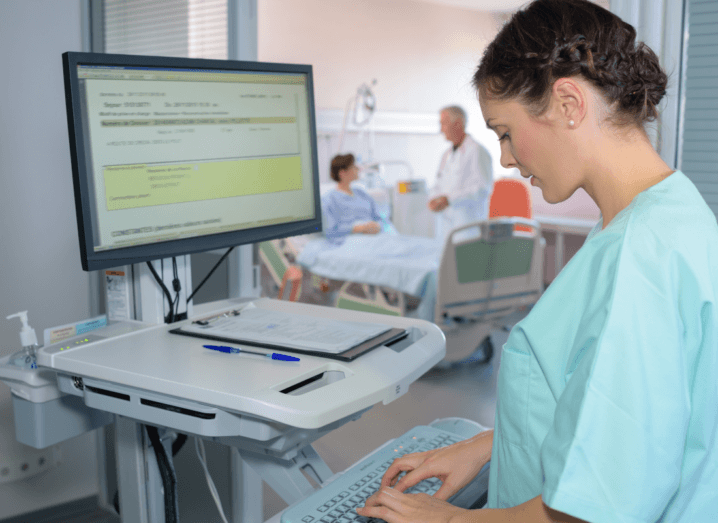 A nurse typing on a computer in a healthcare facility. In the background a doctor deals with a patient lying in a bed.