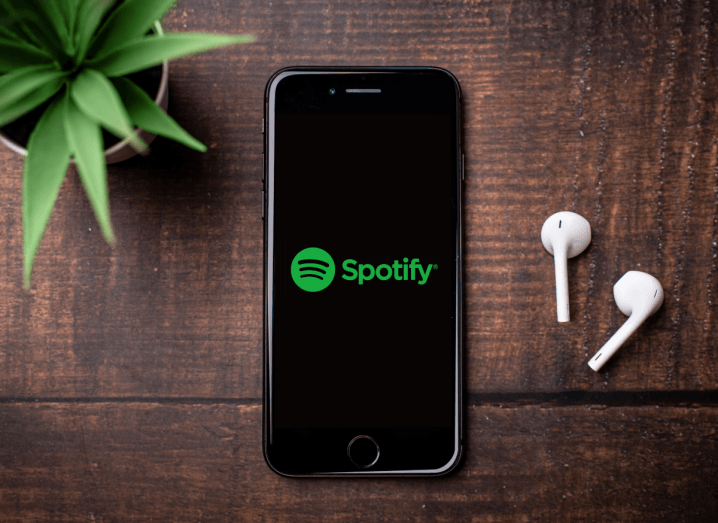 An iPhone screen displaying the Spotify logo. The phone is on a wooden surface beside a potted plant and a pair of AirPods.