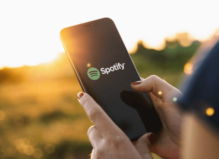The Spotify logo displayed on a phone screen. A woman is holding the phone outdoors, with bushes and grass out of focus in the background.