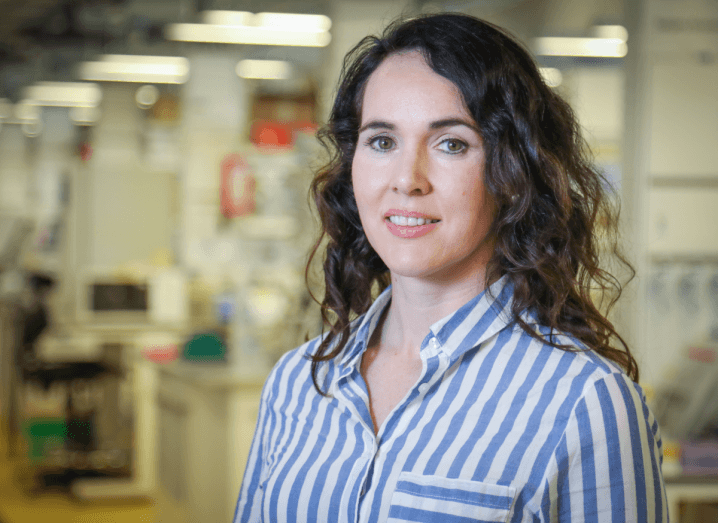 A woman with long, brown, curly hair wearing a blue and white striped blouse stands in a lab smiling.