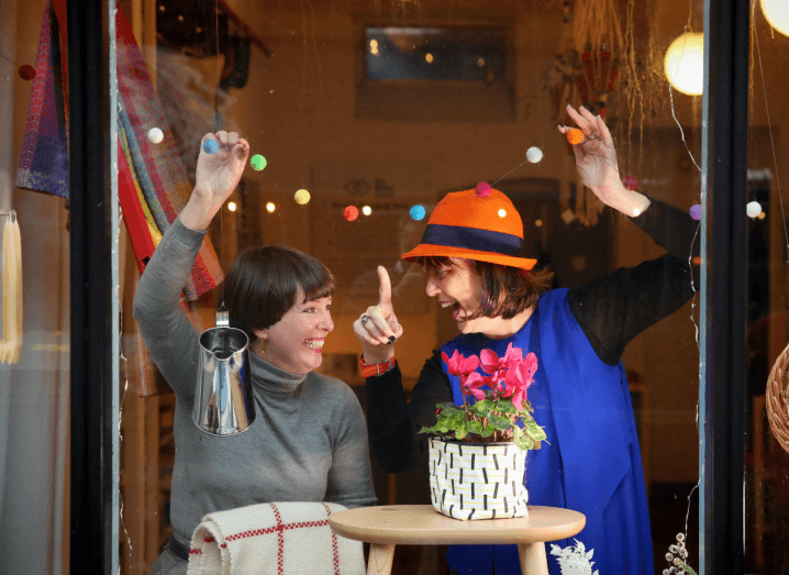 Two women in the window of a shop. One is wearing a grey jumper and the other is wearing a blue dress over a black jumper, with an orange hat.