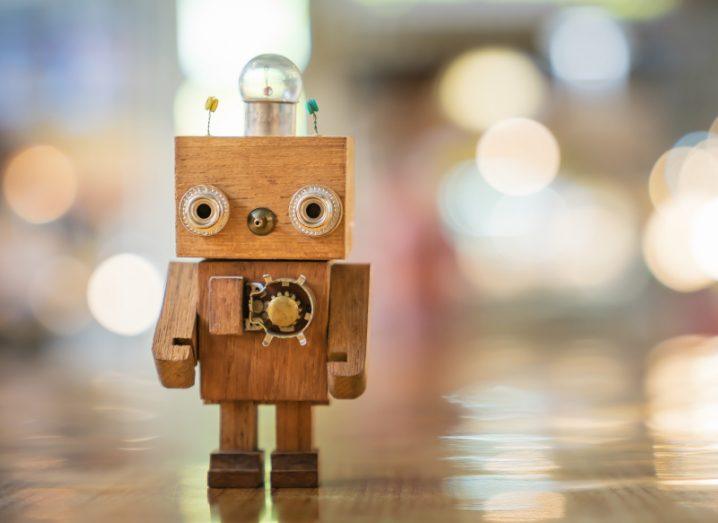 A small, toy wooden robot with a lightbulb on its head stands on a table against a blurred background of lights.