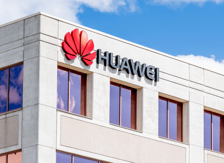 The Huawei logo on an office building under a blue sky with some clouds.