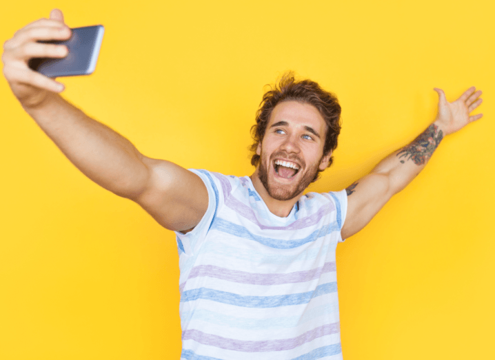 A man taking a selfie in front of a yellow background. He has both arms outstretched and is wearing a striped T-shirt. He has brown hair and a beard.