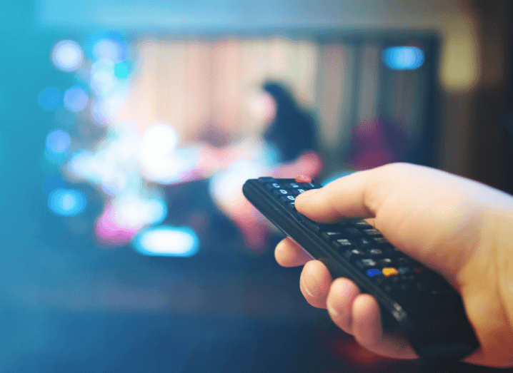 A hand holding a remote control in front of a television. The television screen is out of focus.