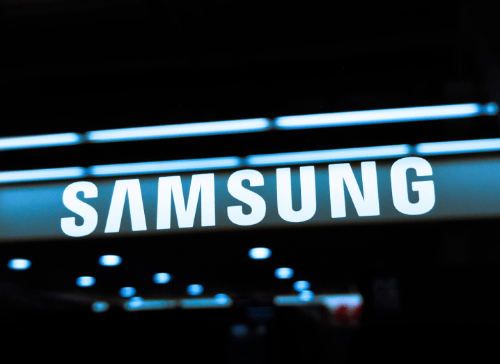 The Samsung logo on a black glass surface, with reflections of lights from a Samsung store dotted underneath the logo.