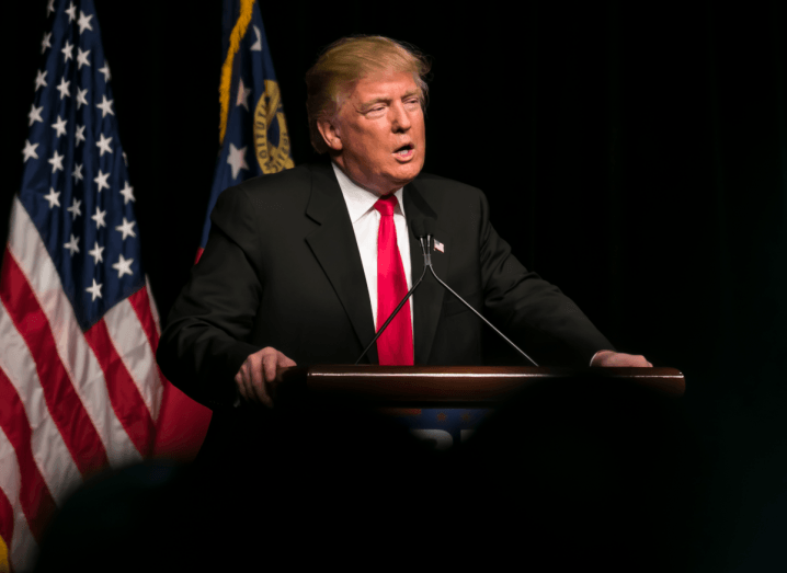 Donald Trump standing at a podium wearing a black suit and a red tie in front of an American flag.