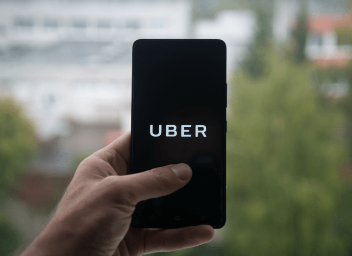The Uber logo displayed on a black screen, held in a hand in front trees and buildings.