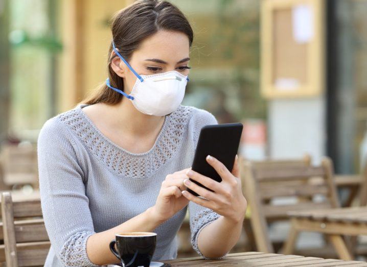 Woman wearing a facemask while sitting at a table with a coffee while looking at a phone.