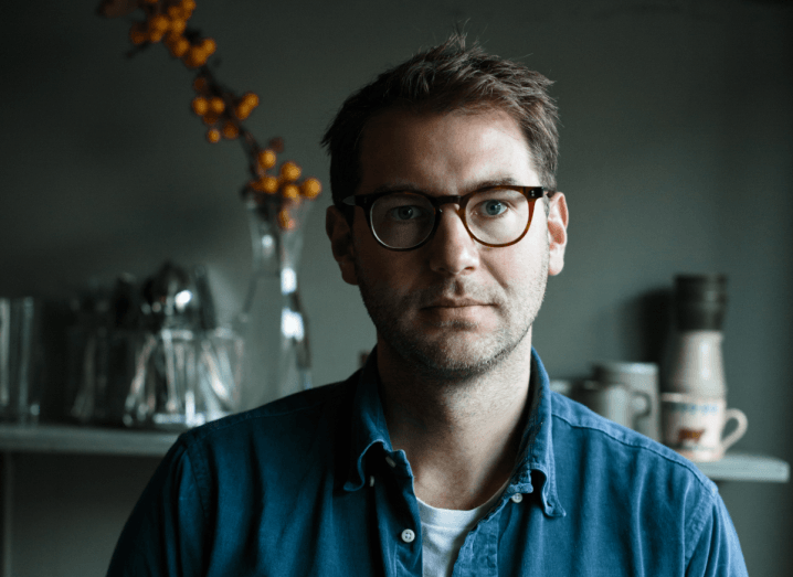 Image of a man wearing glasses and a blue denim shirt sitting in a dark kitchen.