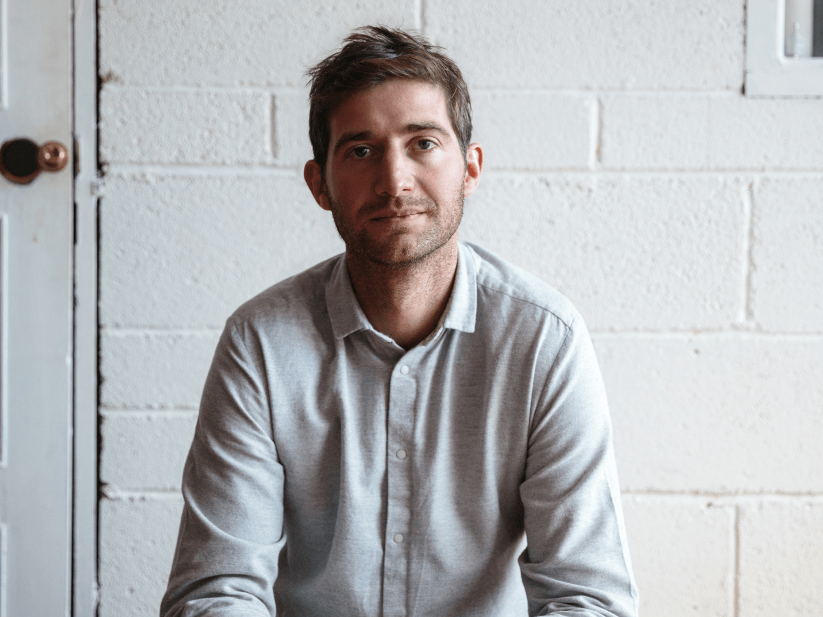 A man in a white shirt sitting in front of a white brick wall. He has brown hair and stubble.