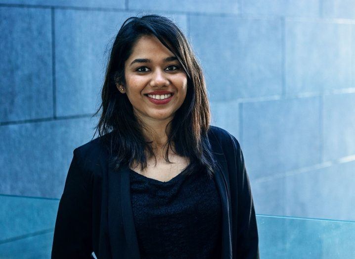 Nidhi Kedia-Mehta smiling in a black top against a blue-brick wall background.