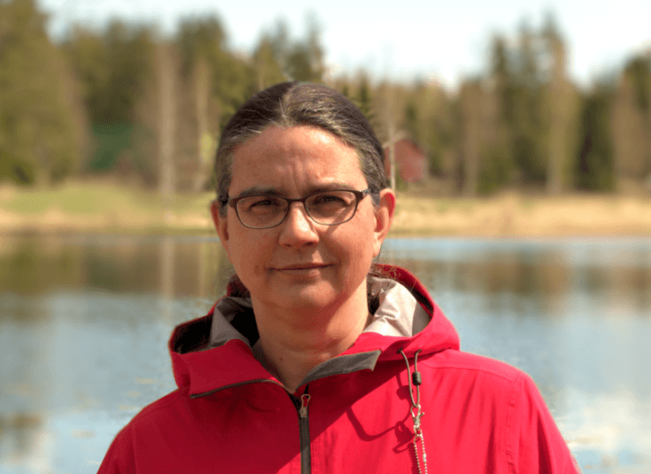 A head and shoulders image of a woman, Dr Silke Holtmanns, wearing a red jacket in front of a picturesque lake.