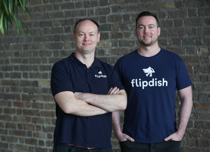 Two men in navy T-shirts with the Flipdish logo stand in front of a brick wall.