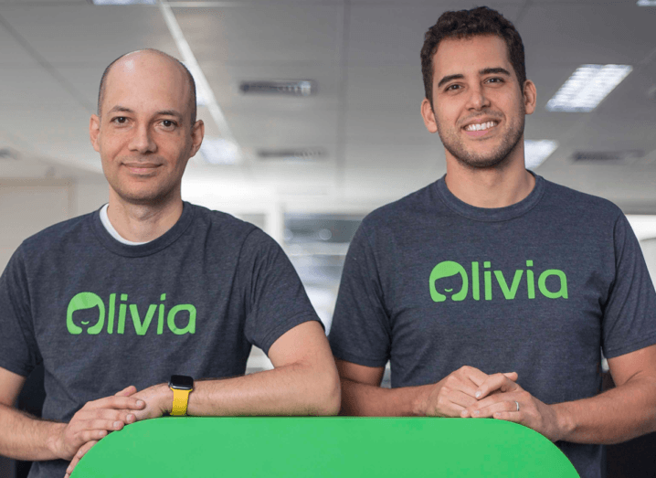 A man with a shaved head and a man with brown hair stand beside each other with their hands on a green surface. They are both wearing grey T-shirts with the logo for Olivia.