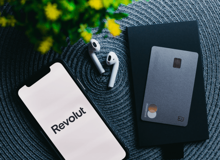 An iPhone on a surface beside a pair of Airpods and a Revolut metal card. The phone's screen displays the Revolut app.