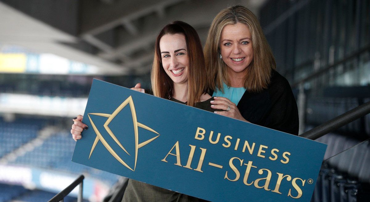 Fiona Descoteaux and Annabelle Conway stand in stadium stalls holding a banner that says Business All-Stars.