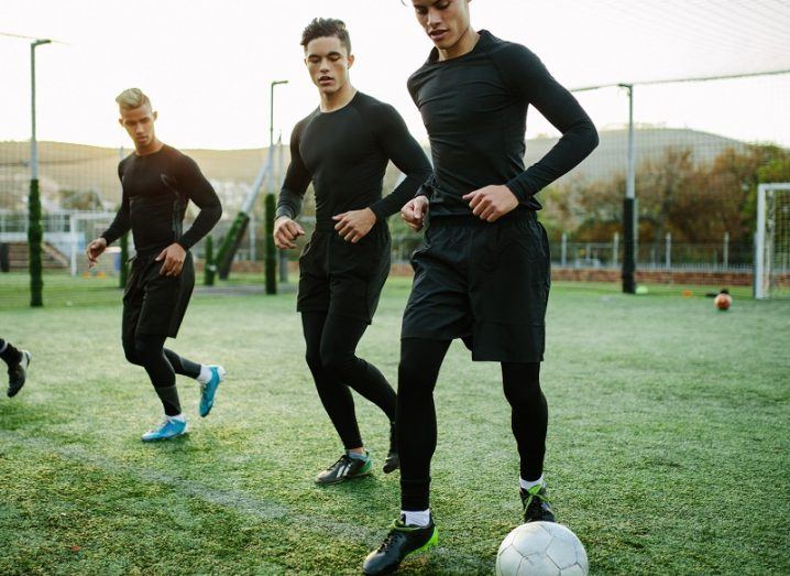 Three football players warming up while wearing all-black sports gear on a football pitch.