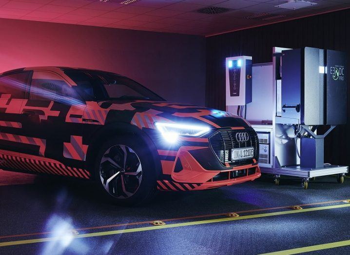 An Audi E-tron car coloured black and neon red, with its lights on beside a bidirectional charging station.