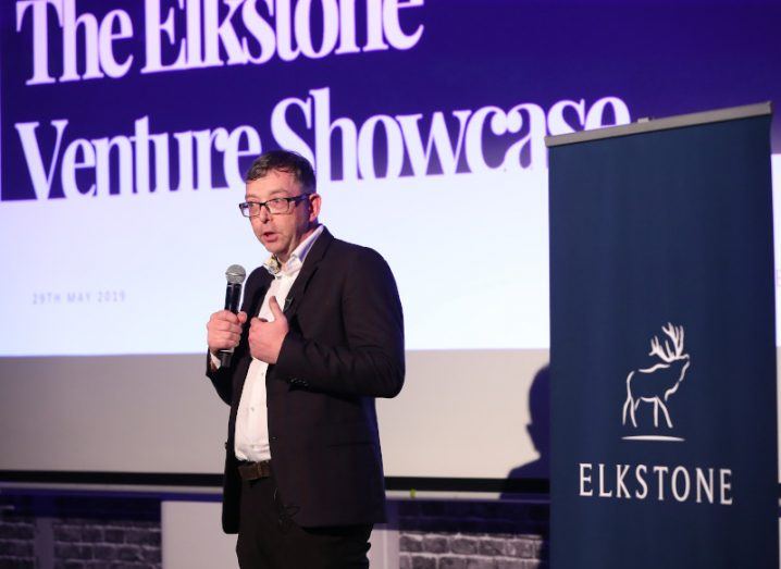 Alan Merriman is wearing a suit and talking into a microphone, while standing in front of a screen that reads 'The Elkstone Venture Showcase'.