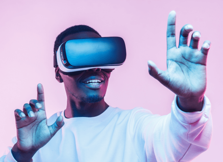A young man in a white jumper wearing a VR headset and standing in front of a pink background. He has his arms raised in front of his body, like he is reaching out for something in virtual reality.
