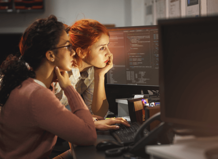 A woman with red hair and a woman with black curly hair work together on a computer with multiple monitors. The woman with the black hair is wearing a pink cardigan and the woman with the red hair is wearing a white blouse.