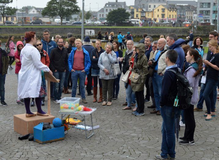 A woman in a lab coat stands on a podium in front of a crowd at Soapbox Science Galway 2019. There are props at her feet.