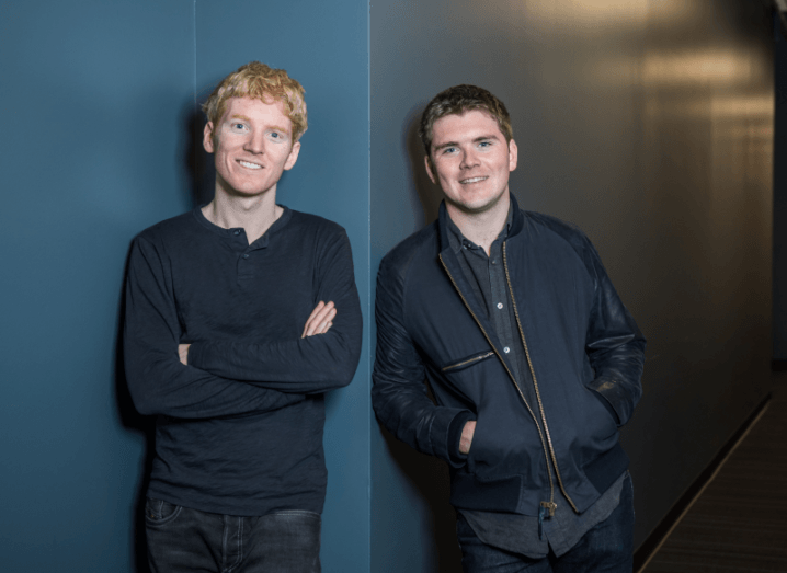 Two young men leaning against a blue wall. The man on the left has red hair and is wearing a navy shirt and the man on the right has brown hair and a navy jacket.